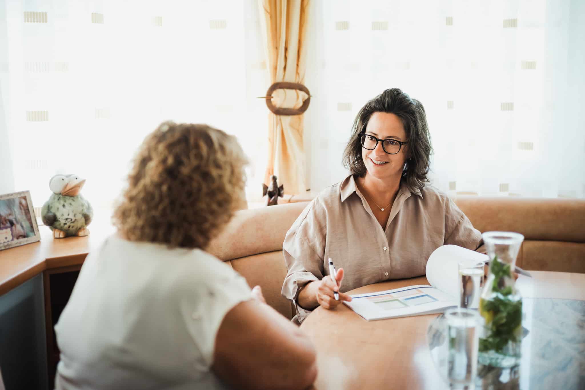 Geschäftsführerin von Nursing on Level und ältere Frau sitzen sich an einem Tisch gegenüber und besprechen gemeinsam Unterlagen bei Tageslicht.