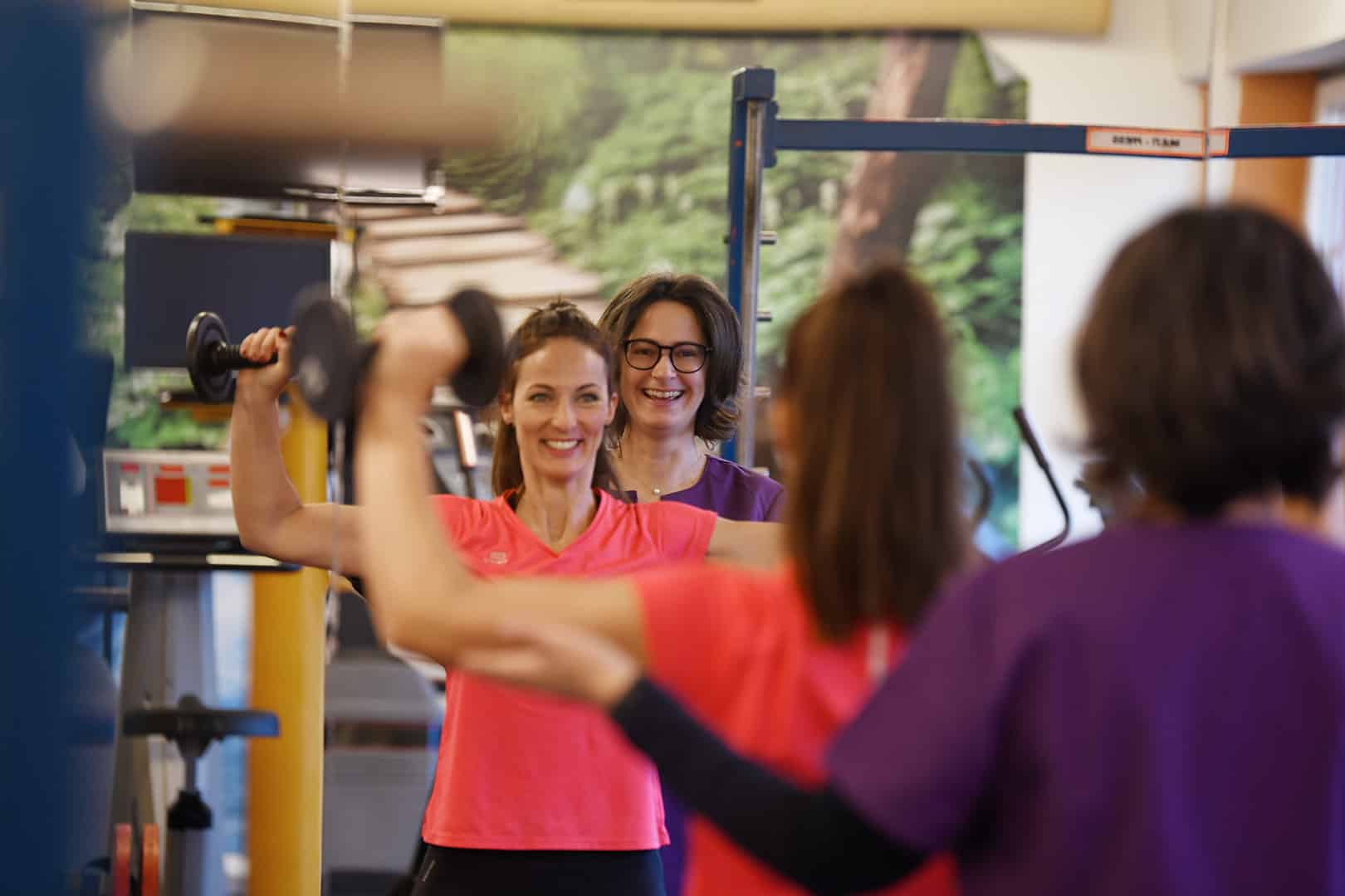Dr. Manuela Sternig bei der Rückbildung. Zwei Frauen sind in einem Fitnessraum, eine davon trägt ein Pinkes T-Shirt, sie hält 2 Hanteln hoch, eine andere steht hinter ihr und unterstützt sie.