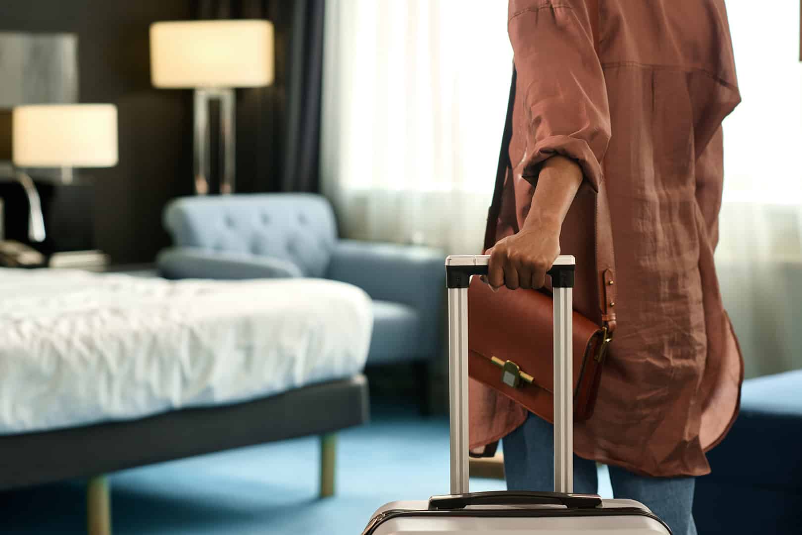 Ein Hotelraum in grau, darin steht eine Person von hinten mit einem Koffer in der Hand.